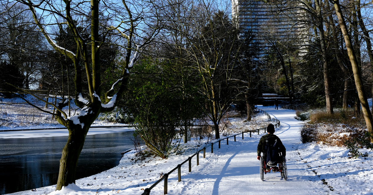 Man in a wheelchair riding down a snowy path in a park by the water