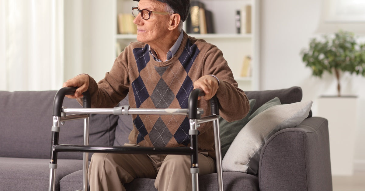 An elderly man in a sweater is sitting on a couch. He has his hands placed on his walker aid in front of him.