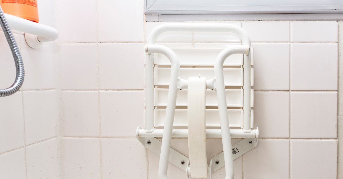 A shower with a folded white shower chair positioned against the wall. There's a grab bar on the other wall of the shower.