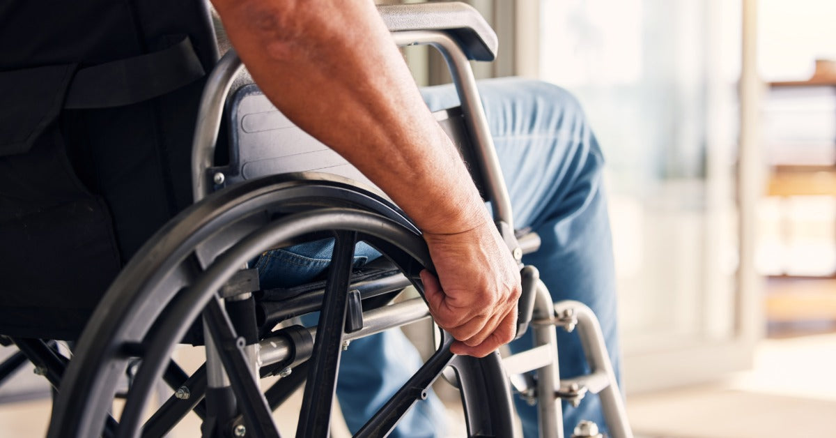 A closeup of a man's hand on the side of wheelchair. He is sitting in front of a window with the sun streaking in.