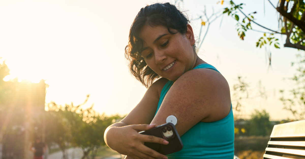 Woman smiling and sitting outside on a bench in the sunshine scanning her CGM with her smartphone