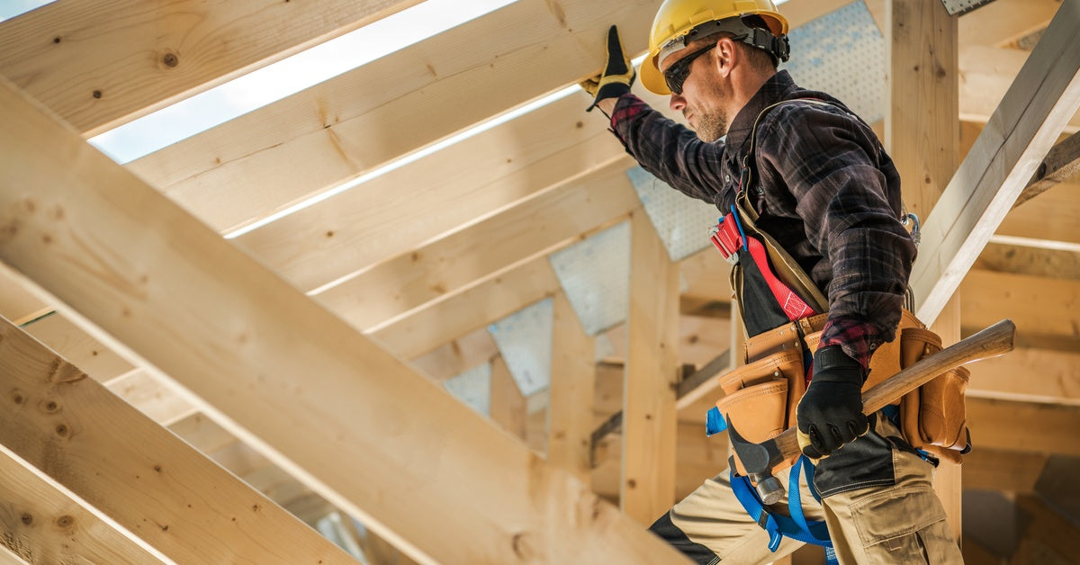 A worker wearing a helmet and with a tool belt looks over a wooden frame of a home, supervising the project.