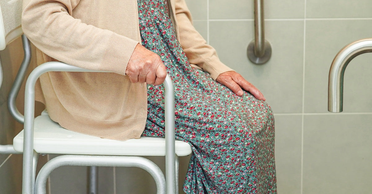 An older woman in a bathroom is sitting in a shower chair and holding the side rail. There are light green tiles in the background.