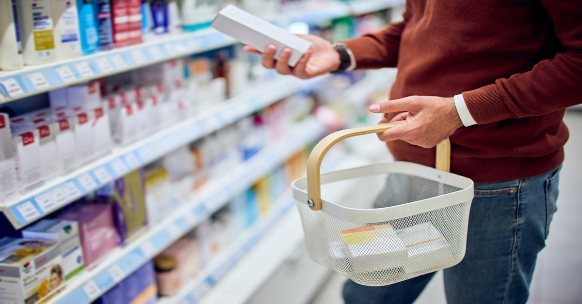 A man shopping in the medicine aisle of a store. He is looking at a box of medicine and holding a shopping basket.