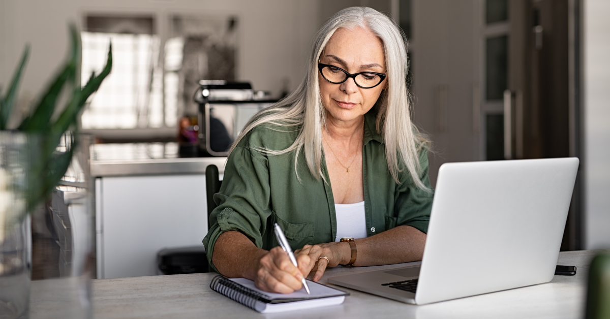 Older woman writing on notepad at the table with her laptop in front of her