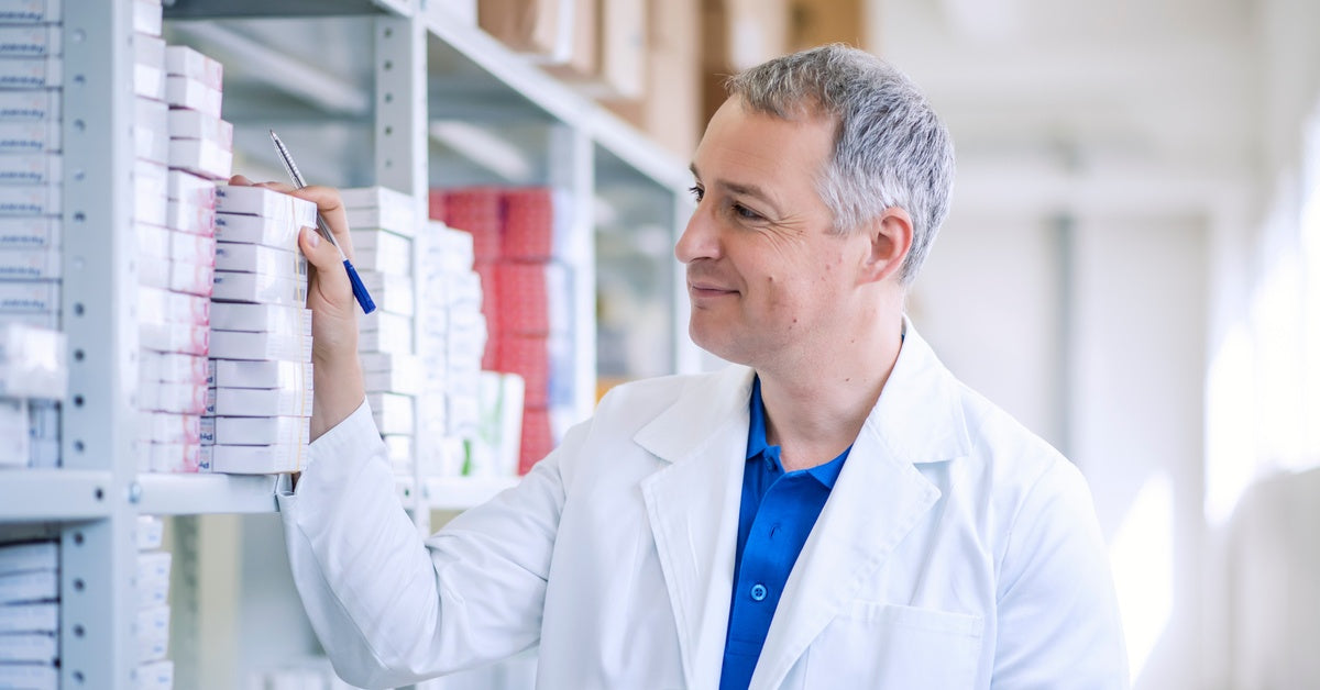A pharmacist working in the backroom and inspecting different shelves with pills while holding a clipboard.