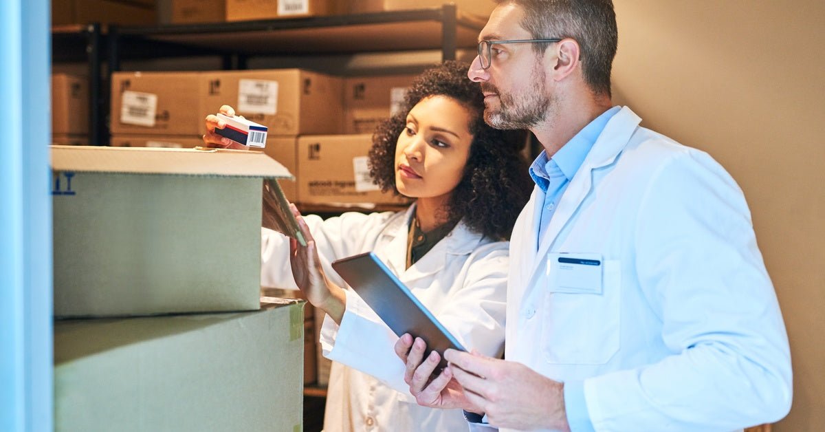 Two doctors in lab coats inspecting a stack of boxes. One of the doctor's is holding a tablet in their hands.