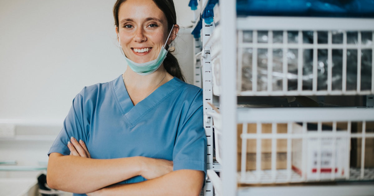 A nurse wearing a face mask is leaning against white, wire rack supply bins in the medical supply closet.
